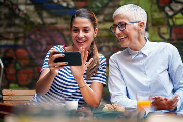 A young beautiful girl is showing a smartphone content to her grandmother while they have a drink in the bar. Leisure, bar, friendship, outdoor