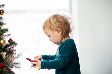 little baby holding a Christmas toy with a Christmas tree in his hands. New year and Christmas at home. Winter Holidays Decorate Christmas Tree