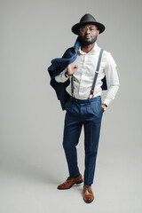 Full-length photo. Attractive stylish african american businessman in a hat and with suspenders holds a jacket with his hand on his shoulder