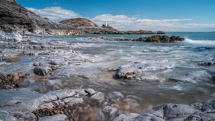 A rocky shoreline, with a white lighthouse on a headland. The beach is Bracelet Bay, Mumbles, south Wales and the day is sunny and clear
