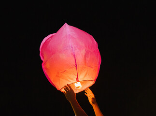 Hands release a paper flashlight into the night sky. Inside the lantern, the fire burns red.