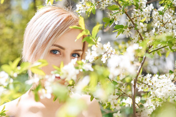 Close up young beautiful blonde woman with blue eyes posing in blooming spring garden with white flowers. Blooming apple or cherry tree. Concept of natural cosmetics, youth, healthy skin and teeth