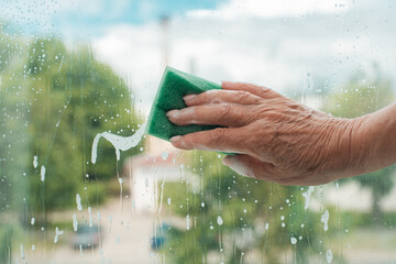 Woman's hand holding a sponge and washing the window, close-up. Hand of a senior woman wiping the...