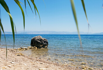 Fototapeta na wymiar Huge stone boulders on Sea coast, huge stone at the seawater and palm tree leafs. Greece summer
