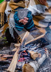 Traveller wrapped a warm plaid holds cup of the hot tea near a bonfire
