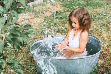 little cute girl in a pink swimsuit flops, having fun and splashing in a basin in the summer in the backyard, the concept of a happy childhood and summer vacation