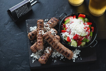 Grilled cevapi or serbian skinless beef sausages with grated bryndza and shopska salad, elevated view on a black stone background, horizontal shot