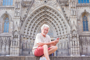 Fototapeta na wymiar Happy man, senior white-haired traveler enjoying visit to cathedral of Barcelona, sitting on staircase holding mobile phone