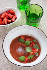 Bowl of tomato gazpacho topped with fresh strawberries, green basil and ice cubes, vertical shot on a beige marble background