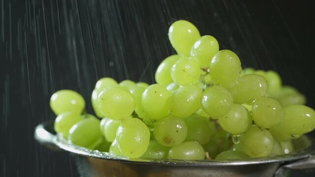 Green grapes in a white plate on a black background close-up. Water is poured onto the grapes, drops flow down over its fruits. The camera moves from left to right, 4K UHD.