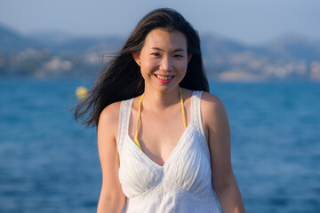 Fototapeta na wymiar young happy and beautiful Asian woman by the sea - Attractive Chinese girl in white dress enjoying relaxed summer holidays at tropical island in travel and lifestyle concept