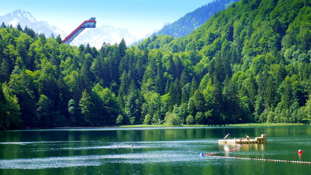 Blick vom Freibergsee in Oberstdorf hinauf zur Skiflugschanze mit gr&uuml;nem Wald unter blauem Himmel