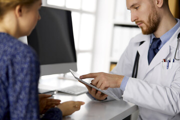 Unknown bearded doctor and patient woman discussing current health examination while sitting and using tablet computer, close-up. Medicine concept