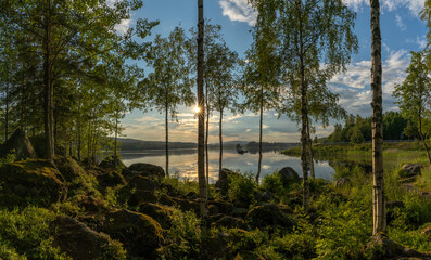 Obraz premium moss covered boulders and birch trees on the shores of a calm idyllic lake in warm evening light and a sun star