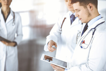 Group of doctors are checking medical names on a computer tablet, with a nurse with a clipboard on the background, while standing together in a sunny hospital office. Physicians ready to examine and