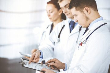 Doctors, two men and a woman, discussing medical exam resoults, while standing at sunny hospital office. Physicians use clipboards for filling up medication history records. Perfect medical service in