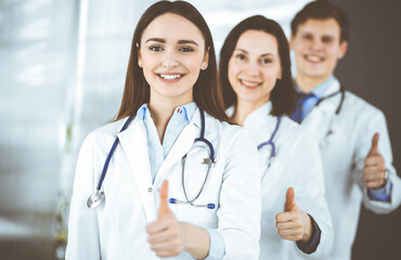 Group of young cheerful doctors is standing as a team with thumbs up in a hospital office and is ready to help patients. Medical help, insurance in health care, best disease treatment and medicine