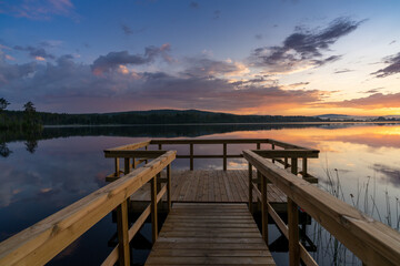 Fototapeta na wymiar wooden pier leads out onto a calm lake with forest on the opposite shore and a colorful sunset sky