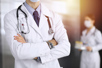 Unknown male doctor in protective mask standing with arms crossed in sunny clinic. Medicine concept during Coronavirus pandemic