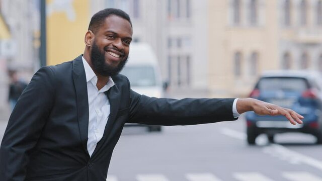 Smiling cheerful benevolent young adult man wearing formal clothes signaling driver to stop, leave by car, joyful guy in suit waiting for taxi standing on sidewalk outdoors. City transport services