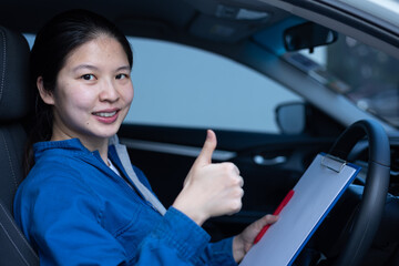 Portrait of pretty, smart Asian woman sitting inside a car, smiling, holding checklist clipboard, putting thumb up. Professional female blue-collar worker or mechanic, and service maintenance concept.