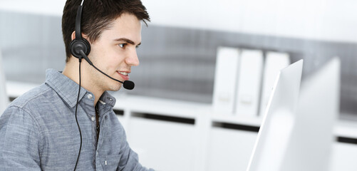 Casual dressed young man using headset and computer while talking with customers online. Call center, business concept