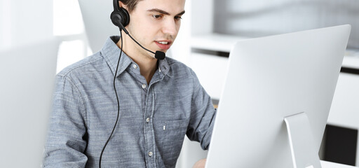 Casual dressed young man using headset and computer while talking with customers online. Call center, business concept