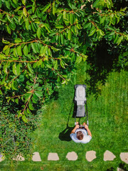 Caucasian gardener mowing backyard garden grass using Cordless electric grass mower. Aerial View. - obrazy, fototapety, plakaty