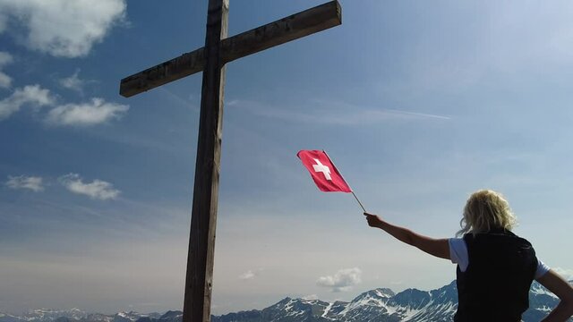 Woman with Swiss flag on top cross of the Piz Scalottas. Plessur Alps in Switzerland in Graubunden canton. Tourist resort for ski activities in winter and mountain bike in summer. SLOW MOTION