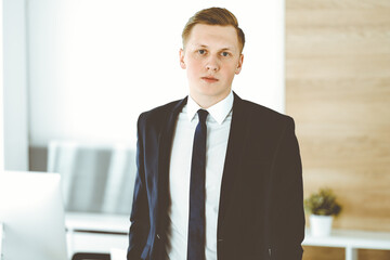 Cheerful businessman standing and looking at camera in office. Headshot of young and successful entrepreneur