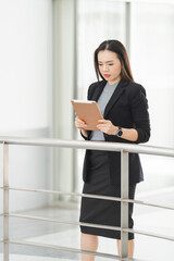 Portrait of a cheerful confident Asian businesswoman in a business suit standing whie using a digital tablet in the business building. Business stock photo