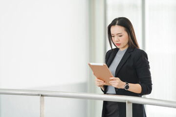 Fototapeta premium Portrait of a cheerful confident Asian businesswoman in a business suit standing whie using a digital tablet in the business building. Business stock photo