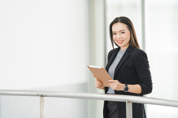 Fototapeta na wymiar Portrait of a cheerful confident Asian businesswoman in a business suit standing whie using a digital tablet in the business building. Business stock photo