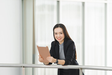 Fototapeta na wymiar Portrait of a cheerful confident Asian businesswoman in a business suit standing whie using a digital tablet in the business building. Business stock photo