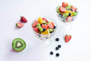 pieces of fresh fruits and berries with whipped cream and chocolate in a transparent bowl on a white table. 
