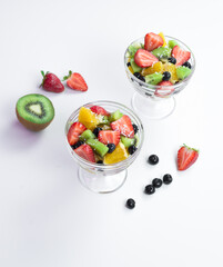 pieces of fresh fruits and berries with whipped cream and chocolate in a transparent bowl on a white table. 