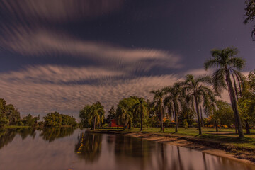 Night shot of a quiet riverside and palm trees in the shore with moving clouds. Stars can be seen...