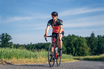 Fototapeta na wymiar Angled photo, of a young woman professional cyclist, riding her road bike, on a paved road amidst nature, illuminated by sunlight. Sport Equality concept.