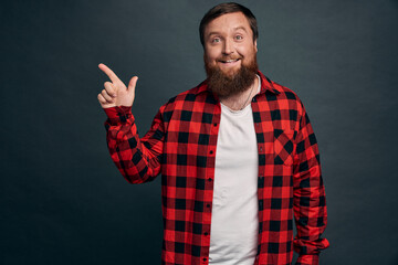Impressed bearded hipster man in red checkered shirt, pointing left and smiling amused, sharing information where find great product, advice check out promo, standing satisfied green-grey background