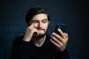 Young confused man looking in smartphone on background of black brick wall.