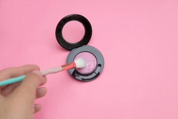 a makeup brush in your hands and pink shiny blush on a pink background copy space