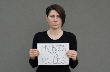 Middle age woman with short black hair looking at camera, holding banner My Body My Rules in front...