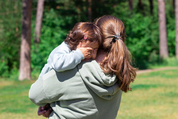 A young woman standing with her back, holding a baby in her arms. A young mother walks with her daughter in a park in nature. A little girl hides her face with hands. Family, love. Sunny day