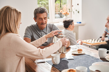 Male pensioner sitting at the table with his caregiver and drinking tea
