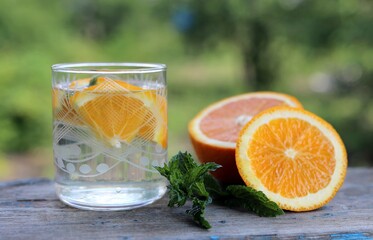 fresh water, orange, green mint leaves in a glass glass with a cut orange on a blurred green bokeh background