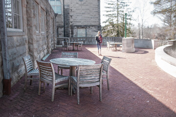 Fototapeta na wymiar Patio at University with wooden tables and chairs in shade in foreground and student with backpack and landscape blurred in distance.