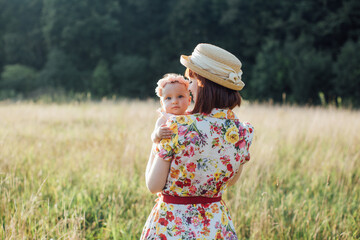 Cute baby girl and mommy are hugging in summer field. Back view of pretty woman mother holding her baby on hands. Little daughter looks at camera. Happy family, mom and daughter on field