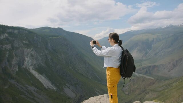 Tourist takes pictures on smartphone camera in mountains. Young woman traveler with backpack shoots video. Travel blogger on hike in mountains against backdrop of clouds