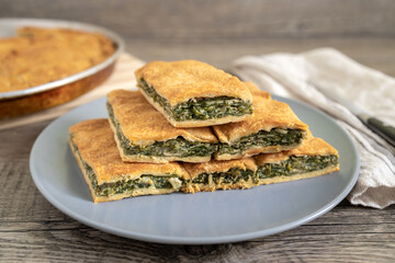 Spinach pie cut in pieces and served on a plate. Greek spanakopita - savory greens pie - on wooden table with pan, kitchen knife and towel.