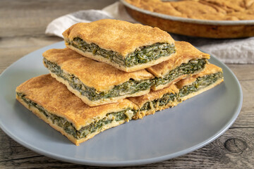 Spinach pie cut in pieces and served on a plate. Greek spanakopita - savory greens pie - on wooden table with pan and towel.